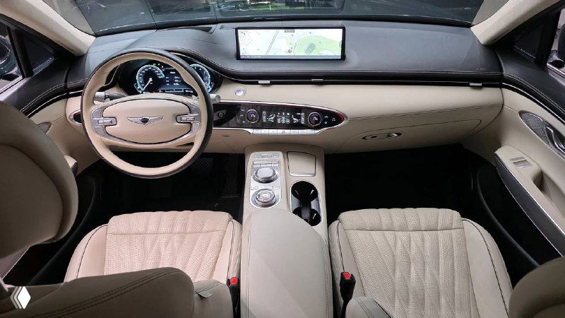Фотография интерьера Genesis GV70 2022: салон светлой бежевой отделки, панель приборов, центральный тоннель и рулевое колесо в кадре.
