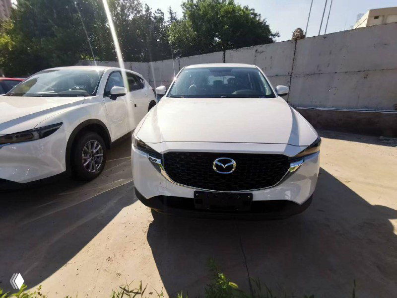 Фронтальный вид белой Mazda CX-5 спереди, видно решетку радиатора и капот; парковочное место у бетонного забора.
