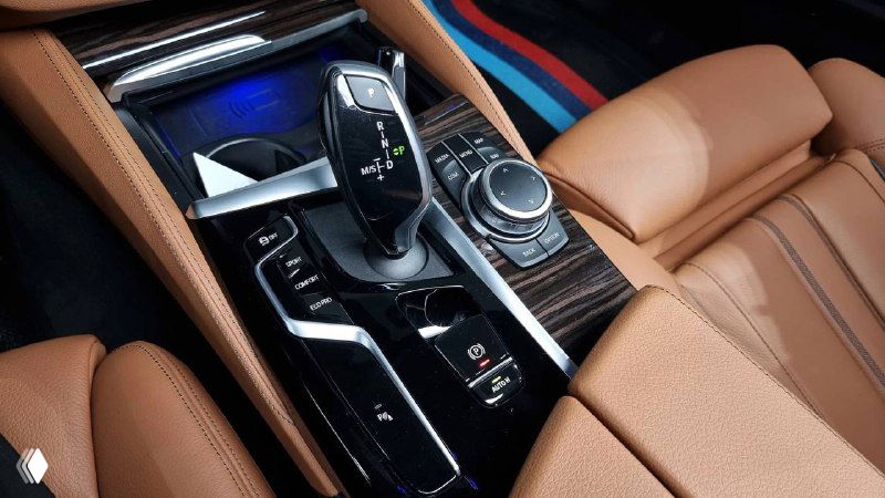 Ближний план центральной консоли и селектора передач BMW 520i M‑Sport: элементы управления, ручка селектора и отделка под дерево/глянец.