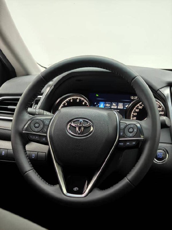 Крупный план руля и приборной панели Toyota Camry 2020, видны кнопки на руле, подсветка приборов и качество отделки салона на демонстрационном фото.