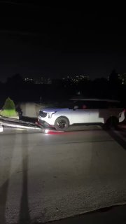 Кадр ночной погрузки: белый внедорожник на пандусе автовоза, городские огни на заднем плане, техническая сцена.