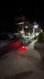 Ночной общий план: несколько автомобилей на автовозе в очереди на погрузку, видны стоп-сигналы и боковые огни платформы.