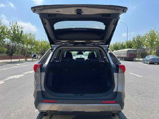 Открытый багажник Toyota RAV4 2021: вид сзади на вместительный грузовой отсек, проезжая часть и городской фон на заднем плане.