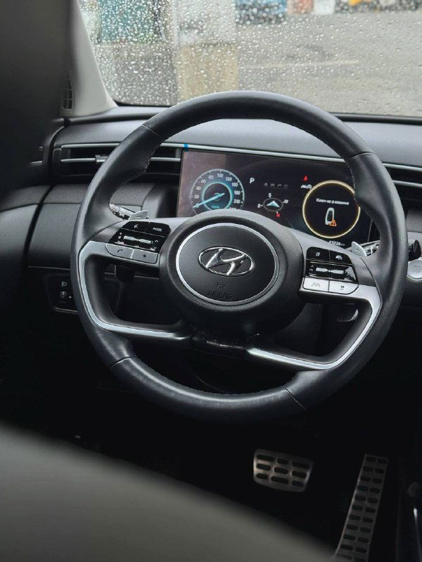 Вид на рулевое колесо и цифровую приборную панель Hyundai Tucson с подсветкой приборов, дождевые капли на лобовом стекле и общая компоновка передней панели.