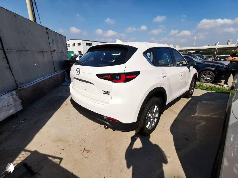 Белая Mazda CX-5, вид сзади-слева: видны задние фонари, крышка багажника и общий профиль кроссовера на бетонной площадке.