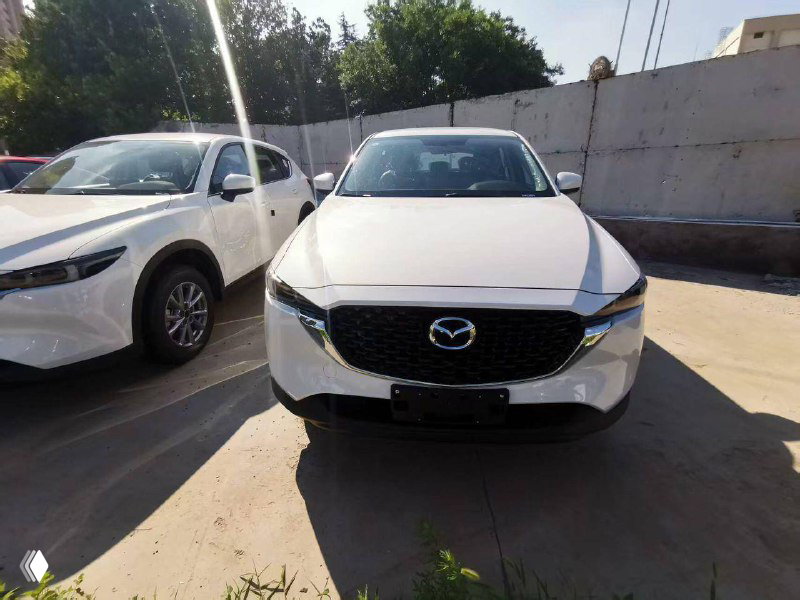 Белая Mazda CX-5 крупным планом спереди, прямой вид на капот и эмблему Mazda в центре решетки радиатора, автомобиль на солнце.