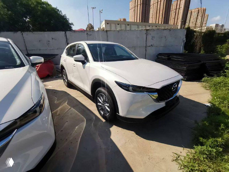 Белая Mazda CX-5 под углом спереди, видно колесные арки и боковую часть кузова; автомобиль припаркован рядом с другими машинами.