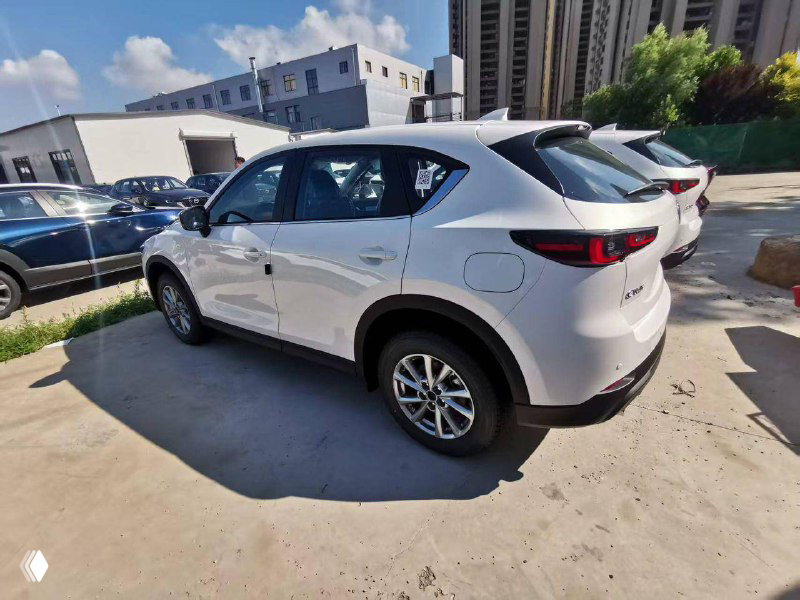 Боковой ракурс белой Mazda CX-5, видно заднюю четверть и профиль двери, автомобиль на бетонной площадке с городскими зданиями на заднем плане.