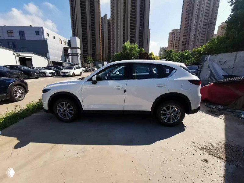 Полный боковой профиль белой Mazda CX-5 на парковке, ровная проекция кузова и дисков колес при дневном свете, окружающая индустриальная зона.