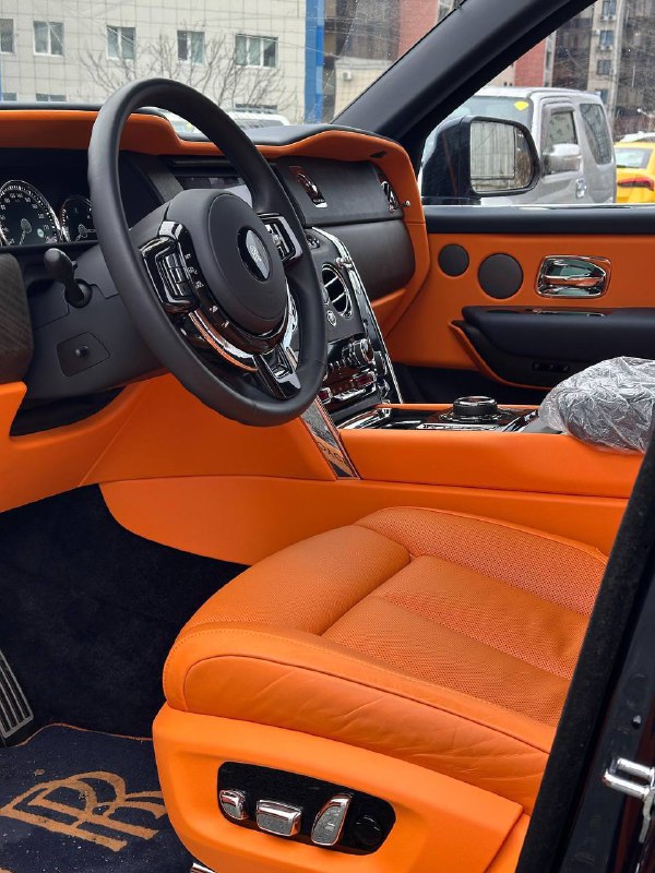 Передняя часть салона Rolls‑Royce Cullinan с яркой оранжевой кожей: руль, водительское сиденье и панель управления рядом с дверью.