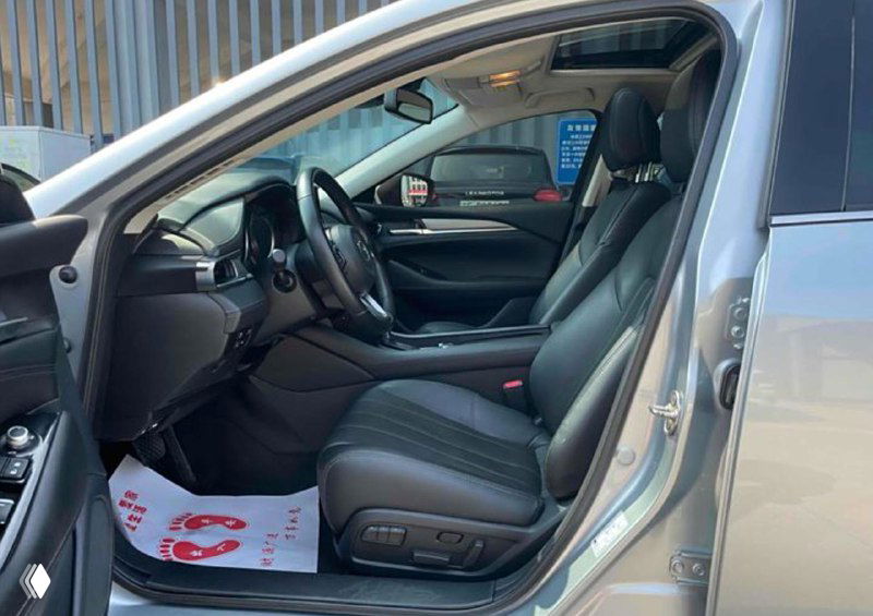 Передний ряд салона Mazda Atenza с тёмной кожаной отделкой сидений, панель приборов и руль на фоне открытой двери, виден коврик и элементы управления