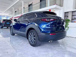 Задняя часть синего Mazda CX-30 2020 в салоне, вид под углом; заметны задние фонари, бампер и открытая демонстрационная зона с плакатами.