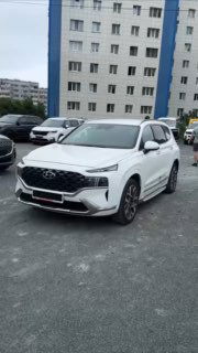 Белый Hyundai Santa Fe в городском паркинге, передний и боковой ракурсы автомобиля на фоне многоквартирного дома — кадр для видеообзора.