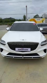 Белый Genesis GV70 2023 года — фронтальный вид автомобиля на открытой стоянке в Южной Корее, превью видеоотчёта о прибытии автомобиля для клиента