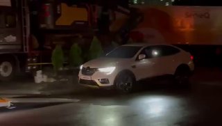 Белый легковой автомобиль вблизи платформы автовоза ночью, включённые фары, рабочая площадка и грузовой транспорт на заднем плане.