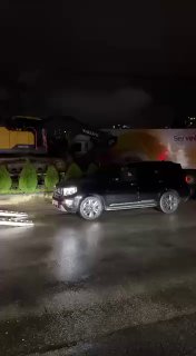 Чёрный внедорожник на дороге у стоящих автовозов в темноте, подсветка фар и отражения на мокром асфальте, ночная логистика грузов.