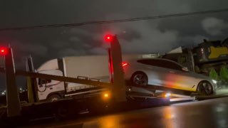 Автомобиль заезжает на платформу автовоза по аппарели ночью; заметны боковые огни грузового транспорта и мокрая дорога.