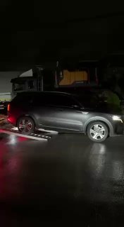 Серый кроссовер на платформе автовоза в тёмное время суток, видна часть грузовика и отражение света на корпусе машины.