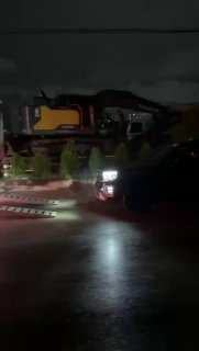 Ночной кадр: фронтальная часть грузового транспорта и припаркованные автомобили на платформе, городские огни вдали и влажная поверхность дороги.