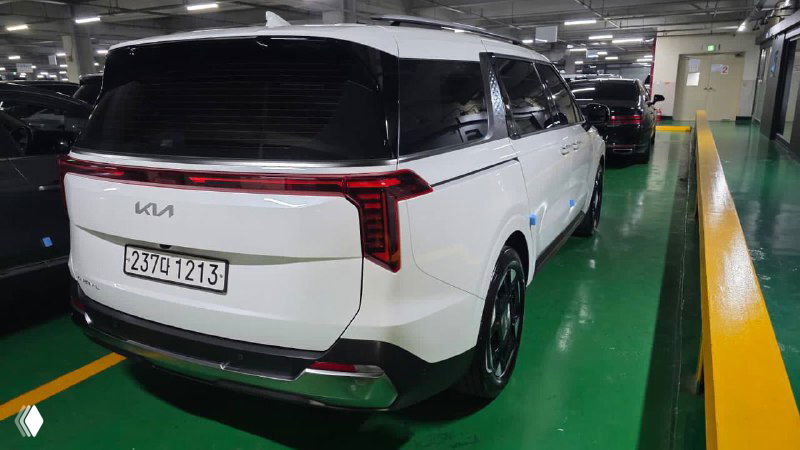 Задняя часть белого KIA Carnival 2024 в крытой парковке: вид на корму, задние фонари, багажную дверь и бампер с регистрационным номером.