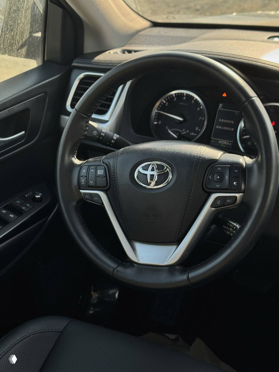 Рулевое колесо Toyota Highlander с логотипом Toyota, вид панели приборов и элементов управления, интерьер в чёрной отделке, крупный план салона.