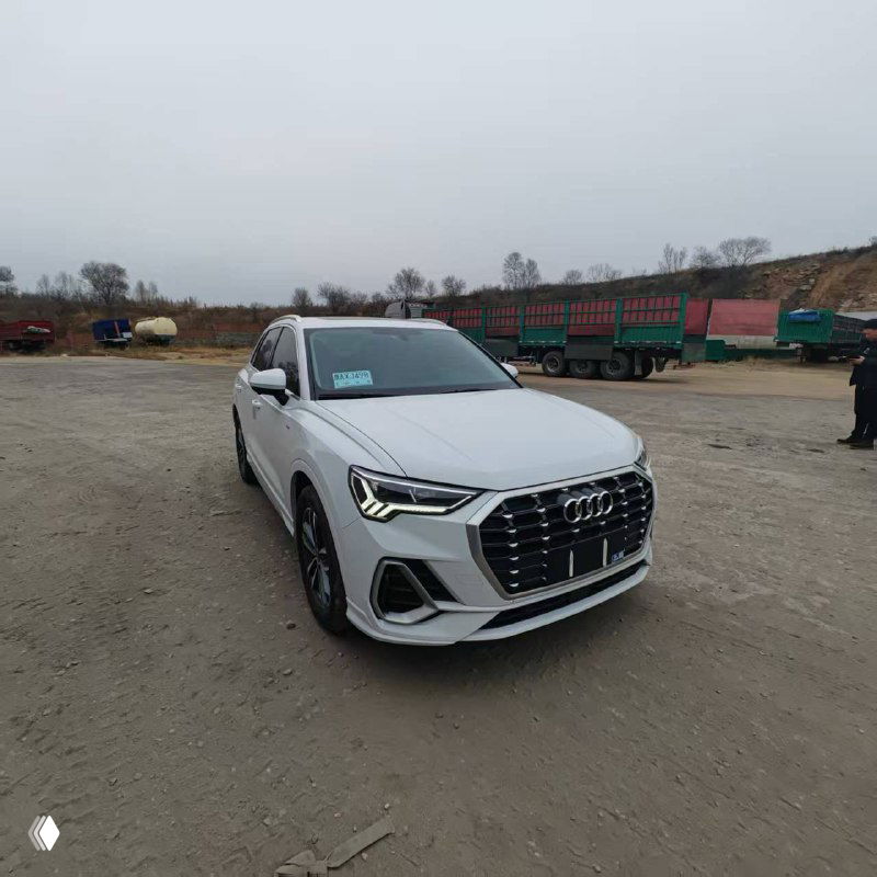 Белый Audi Q3 2022 года, спередний ракурс на стоянке у промышленного склада; видна решётка радиатора, грузовые машины и ограждение на заднем фоне.