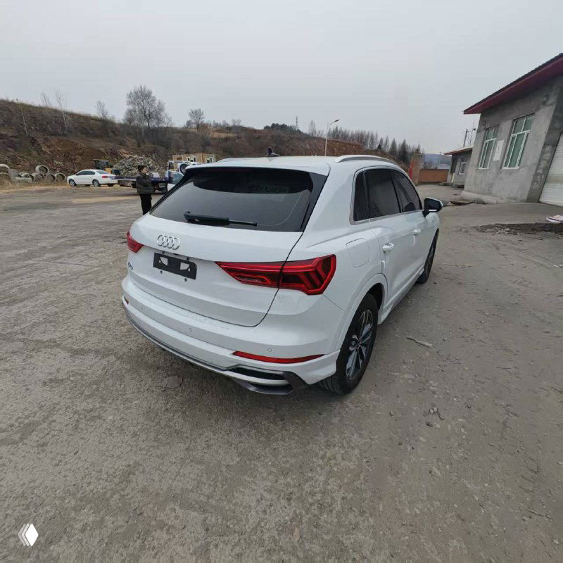 Audi Q3, вид сзади: белый автомобиль у склада, задний бампер и фонарь в фокусе, рядом промзона, грузовой двор и техника на фоне.