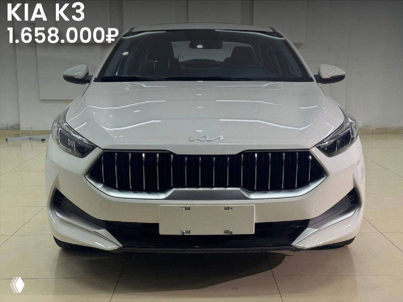 Фронтальный кадр KIA K3 (2021) белого цвета в выставочном зале, видны решётка радиатора, капот и номерной знак.