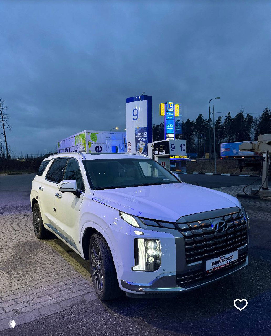 Hyundai Palisade 2023 в белом цвете на стоянке компании при сумерках, вид подчёркивает форму кузова, колёса и включённые фары автомобиля