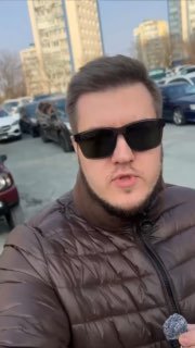 Мужчина в солнцезащитных очках на автостоянке в кадре; на заднем плане припаркованные автомобили и многоэтажные дома в городе.