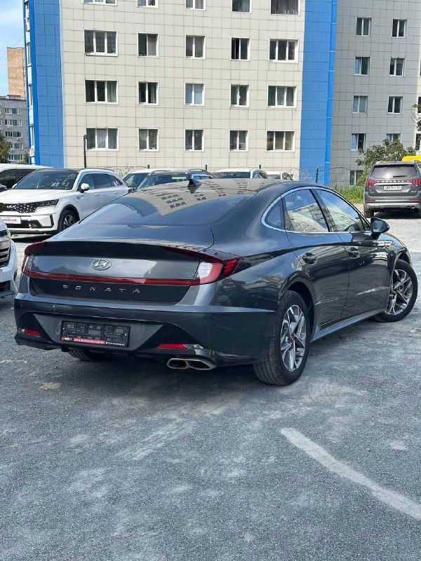 Задняя часть Hyundai Sonata 2022: вид сбоку и сзади, задние фонари и выхлопные патрубки на фоне офисного здания и других автомобилей.