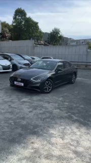 Чёрная Hyundai Sonata на стоянке, вид спереди‑сбоку; автомобиль на асфальтовой площадке с другими машинами и бетонным забором.