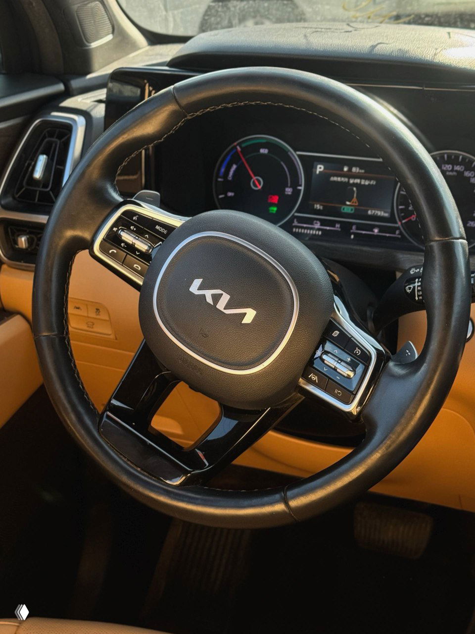 Крупный план многофункционального руля с логотипом KIA и частью приборной панели; видна комбинация приборов и элементы управления на руке, салон бежевого цвета.
