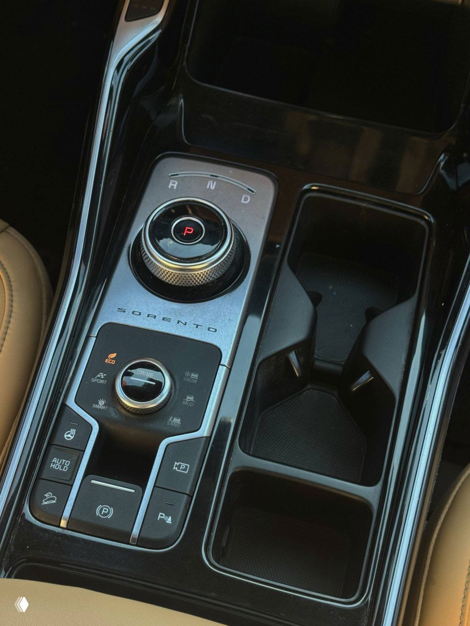 Ближний план центрального туннеля: поворотный селектор передач с маркировкой R N D и кнопками режимов, надпись SORENTO и глянцевая черная отделка рядом с подстаканниками.