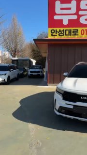 KIA Sorento 2023 белого цвета на открытой парковке в Южной Корее, вид сбоку и часть фасада автосалона на заднем плане