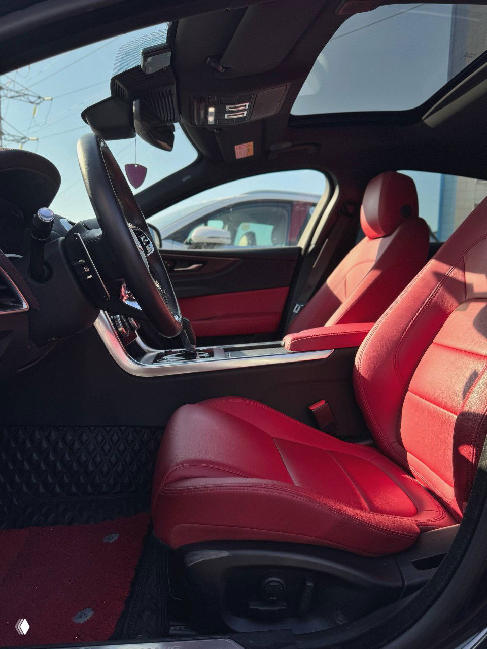 Передние сиденья и панорамный люк Jaguar XE L; вид с открытой двери показывает красную кожаную отделку салона и рулевую колонку.