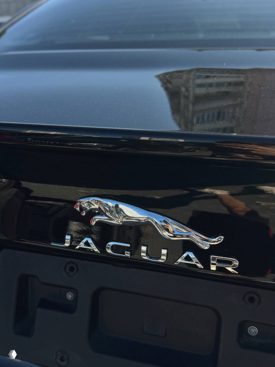 Ближе к эмблеме на крышке багажника — хромированная фигурка и надпись JAGUAR на чёрном лаке кузова, видны отражения окружения.