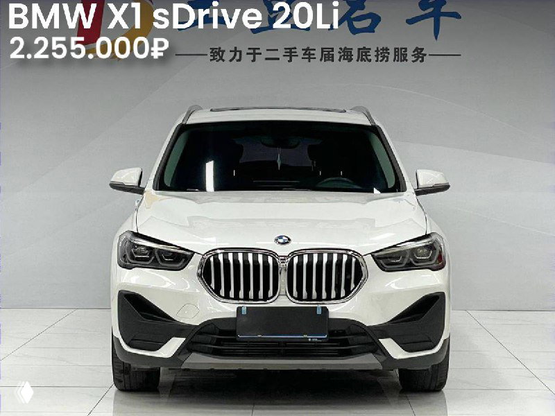 Фронтальный кадр белого BMW X1 sDrive 20Li в салоне: видна фирменная решётка радиатора, цена 2.255.000₽ и отсутствие окраса на кузове.
