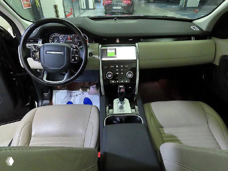 Интерьер Land Rover Discovery Sport 2020: передние сиденья бежевого цвета, панель приборов и руль в салоне на стоянке автосалона.