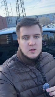 Видео со стоянки: ведущий в тёмной куртке держит микрофон у авто, на фоне видны припаркованные машины, контейнеры и опоры.