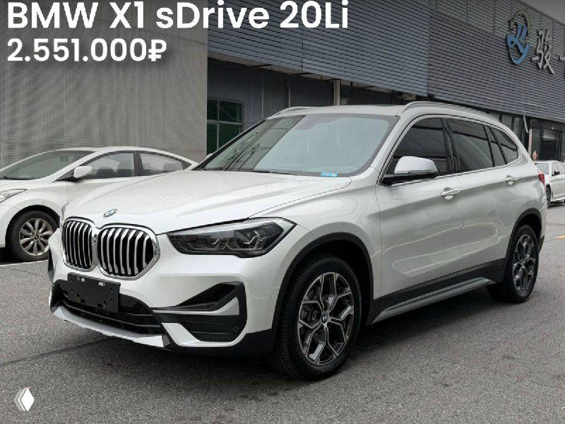 Боковой и фронтальный ракурс BMW X1 sDrive 20Li 2021 года на выставочной площадке; белый кузов, Premium комплектация, цена 2.551.000₽, видны стенды дилера.