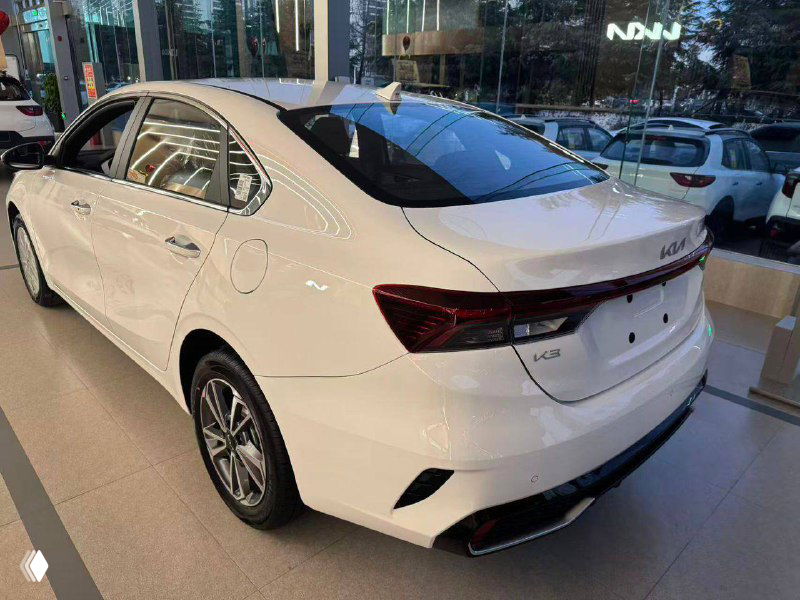 Белый KIA K3 — вид сзади и правого бока в салоне, показаны задние фонари, бампер и общий профиль новой модели на выставке.