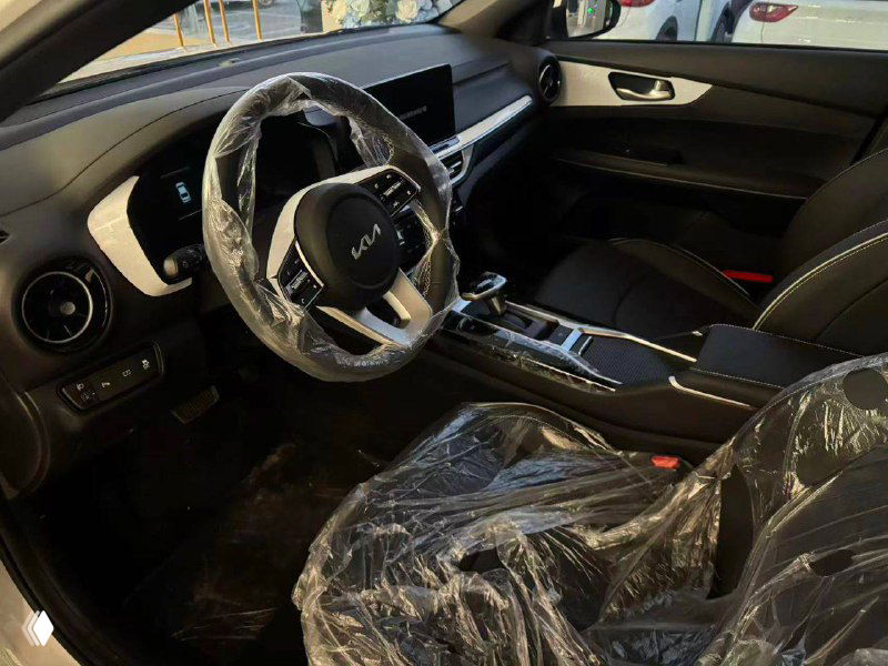 Передняя часть салона и руль KIA K3 — центральная консоль, селектор и сиденья в защитной плёнке, показаны элементы управления и отделка.