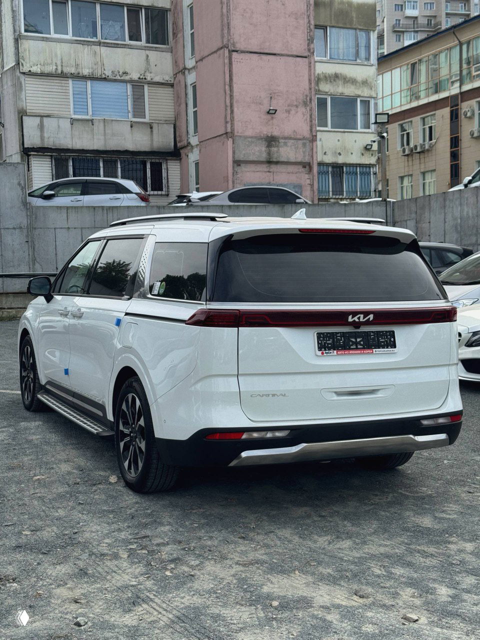 KIA Carnival 2022 вид сзади-по диагонали — багажник, светодиодная задняя оптика и логотип KIA, автомобиль припаркован у жилого дома.