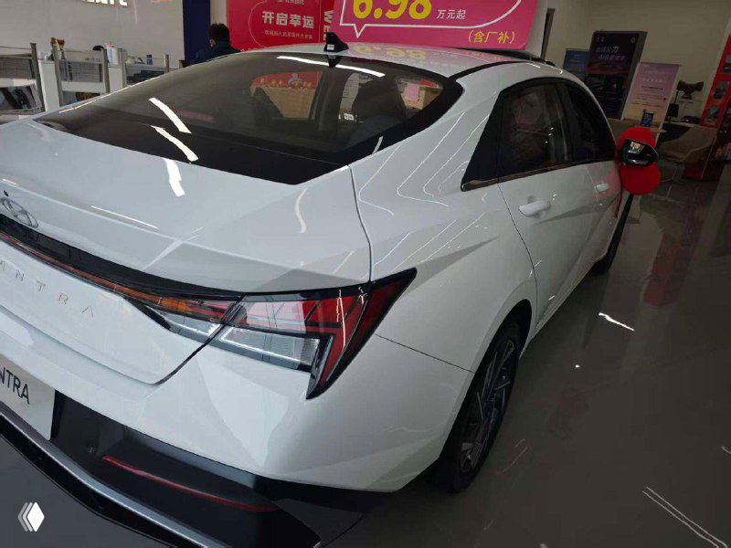 Боковой и задний ракурс белой Hyundai Elantra в выставочном зале: заметны характерные задние фонари и профиль кузова автомобиля.