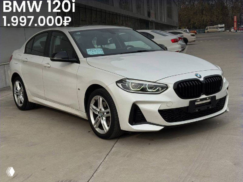 Белый BMW 120i на открытой площадке, вид сбоку‑спереди; подпись с ценой 1.997.000₽ и информационные стикеры на лобовом стекле.