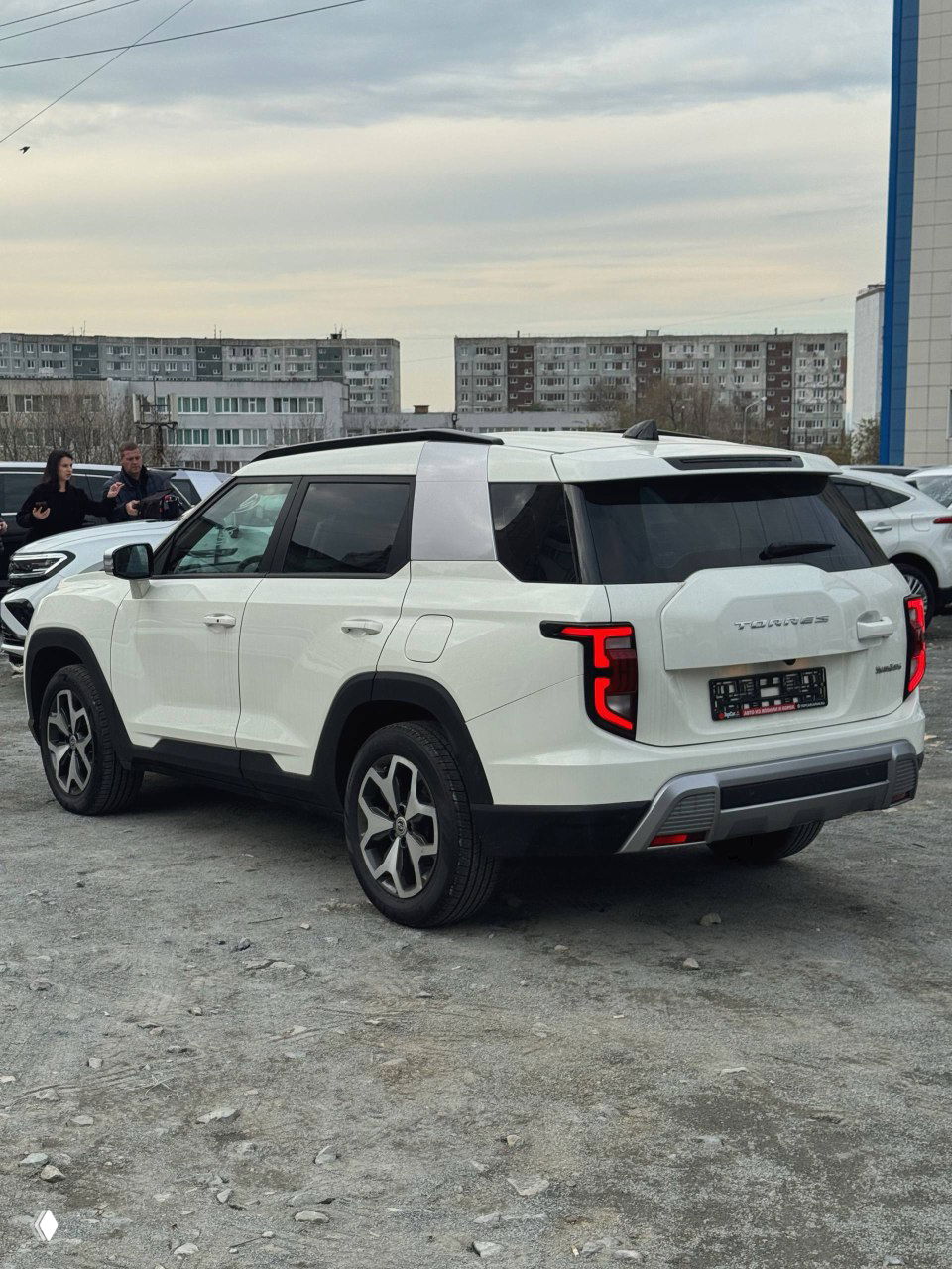 Задняя трёхчетвертная проекция белого SsangYong Torres с характерными задними фонарями и багажной дверью на фоне жилых домов.