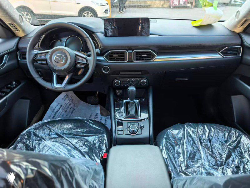 Панель приборов и центральная консоль Mazda CX-5 2025: руль, мультимедийный экран, селектор коробки передач и пластиковая защитная упаковка на сиденьях.