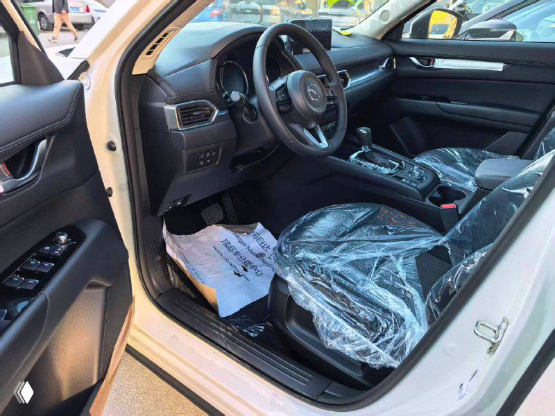 Вид на переднее сиденье и водительскую дверцу Mazda CX-5 2025: рулевая колонка, двери, элементы управления и заводская упаковка на обивке.