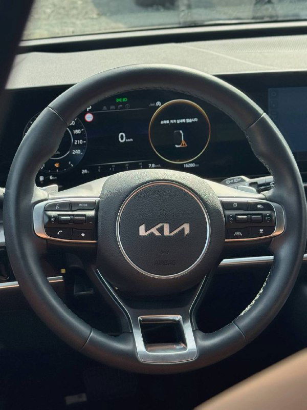 Крупный план рулевого колеса KIA Sportage с новым логотипом KIA и панелью приборов; видно цифровую приборную панель и элементы управления на руле.